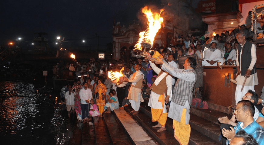 Saffron-clad Hindu priests holding large, multi-tiered brass oil lamps performing the grand Ganga Aarti ritual on the steps of Dashashwamedh Ghat at night, with crowds of people watching from boats.