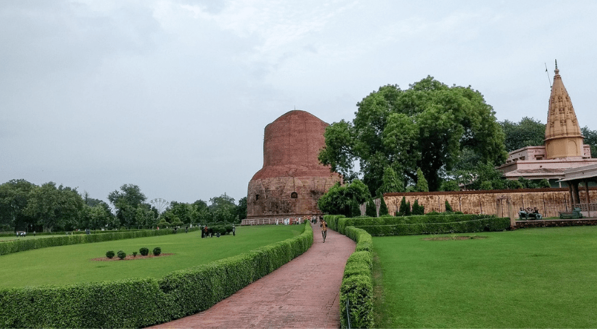 The massive, ancient cylindrical stone Dhamekh Stupa stands tall under a clear sky, surrounded by green lawns and archaeological ruins in Sarnath.