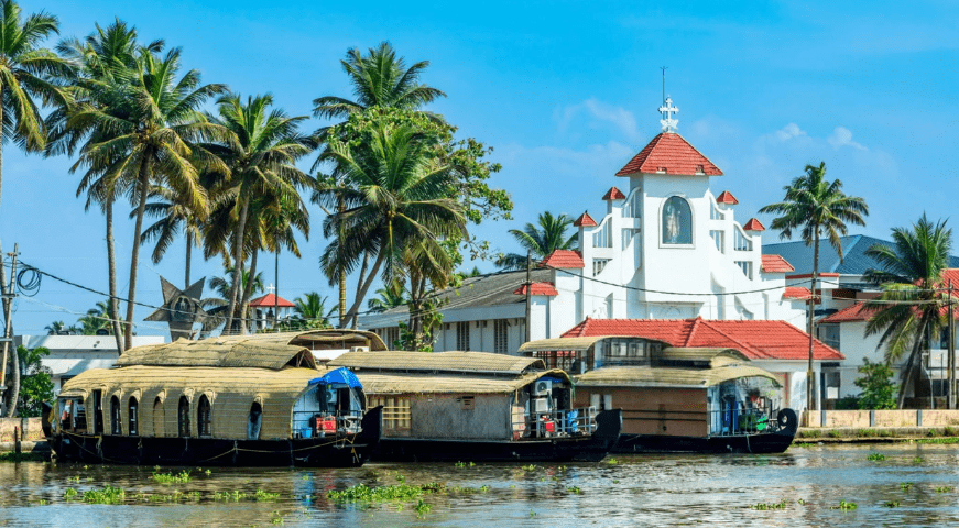A traditional wooden houseboat cruising gently along the calm, palm-fringed backwaters of Alleppey under a clear sky in Kerala.