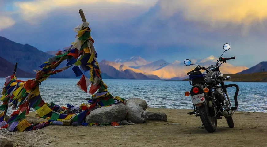 A panoramic view of the snow-covered Khardung La Pass in Ladakh with colorful Tibetan prayer flags fluttering against high Himalayan mountain peaks and a clear blue sky.