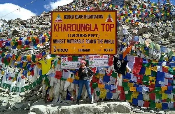 The iconic yellow Border Roads Organisation signboard at Khardungla Top marking the high-altitude pass, heavily surrounded by thousands of colorful Tibetan prayer flags against a rocky mountain peak and a bright blue sky.