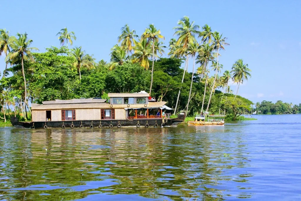 A traditional wooden thatch-roofed luxury houseboat cruising on the calm blue waters of Alleppey backwaters, lined with lush green coconut palm trees under a clear sky.