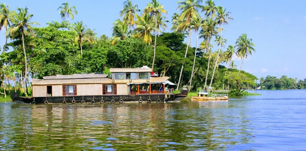 A traditional wooden thatch-roofed luxury houseboat cruising on the calm blue waters of Alleppey backwaters, lined with lush green coconut palm trees under a clear sky.