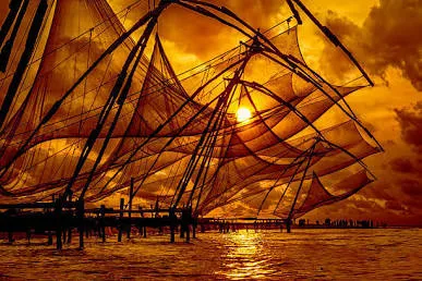 Silhouettes of massive wooden Chinese fishing nets stretched out over the water against a dramatic, glowing orange sunset sky in Fort Kochi, Kerala.